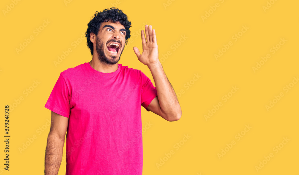 Handsome young man with curly hair and bear wearing casual pink tshirt shouting and screaming loud to side with hand on mouth. communication concept. Handsome young man with curly hair and bear wearing casual pink tshirt shouting and screaming loud to side with hand on mouth. communication concept.