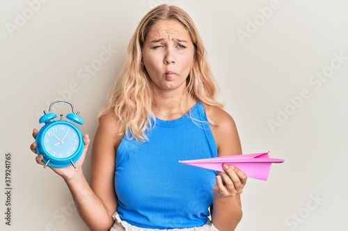 Young blonde girl holding paper plane and alarm clock making fish face with mouth and squinting eyes  crazy and comical.