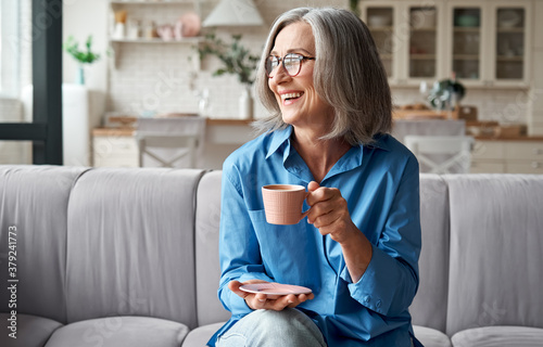 Happy beautiful relaxed mature older adult grey-haired woman drinking coffee relaxing on sofa at home. Smiling stylish middle aged 60s lady enjoying resting sitting on couch in modern living room.