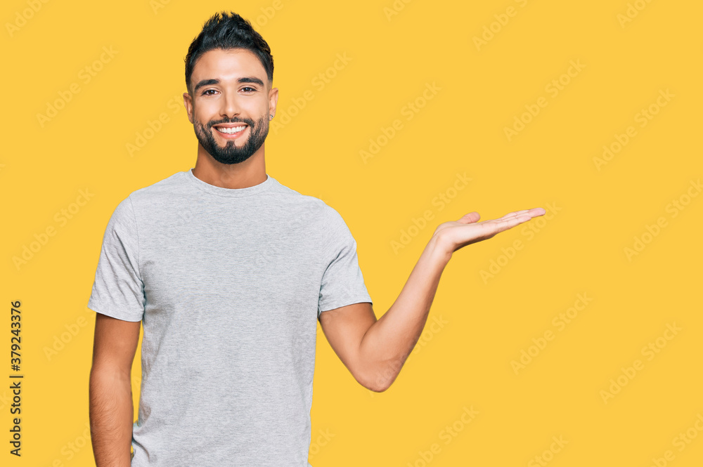 Young man with beard wearing casual grey tshirt smiling cheerful presenting and pointing with palm of hand looking at the camera. Young man with beard wearing casual grey tshirt smiling cheerful presenting and pointing with palm of hand looking at the camera.