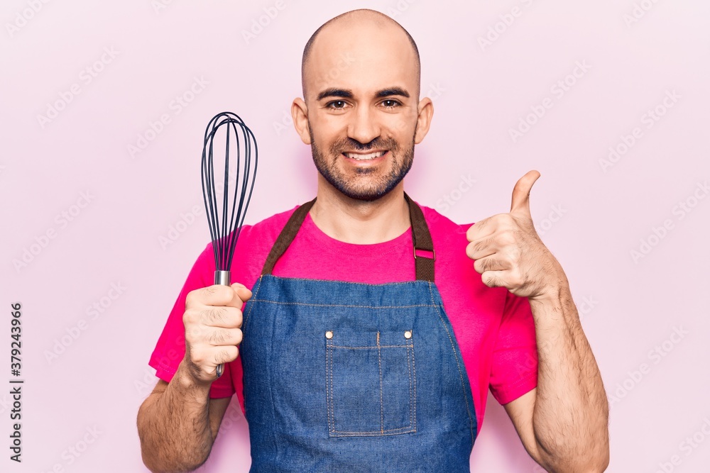 Young handsome bald man wearing apron holding whisk smiling happy and positive, thumb up doing excellent and approval sign Young handsome bald man wearing apron holding whisk smiling happy and positive, thumb up doing excellent and approval sign