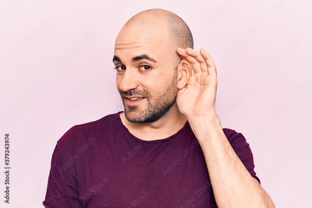 Young handsome bald man wearing casual t shirt smiling with hand over ear listening and hearing to rumor or gossip. deafness concept.
