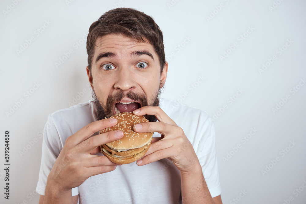 A man eating a hamburger on a light background in a white T-shirt cropped view close-up hunger fast food