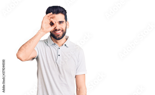 Young handsome man with beard wearing casual polo doing ok gesture with hand smiling, eye looking through fingers with happy face.