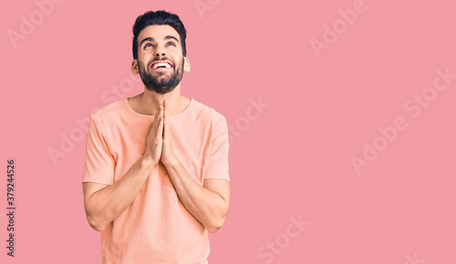 Young handsome man with beard wearing casual t-shirt begging and praying with hands together with hope expression on face very emotional and worried. begging.