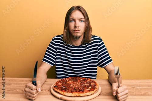 Handsome caucasian man with long hair eating tasty pepperoni pizza relaxed with serious expression on face. simple and natural looking at the camera.