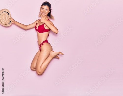 Young beautiful girl on vacation wearing bikini smiling happy. Jumping with smile on face holding summer hat doing ok sign with thumb up over isolated pink background