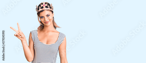 Young beautiful woman wearing king crown smiling with happy face winking at the camera doing victory sign. number two.