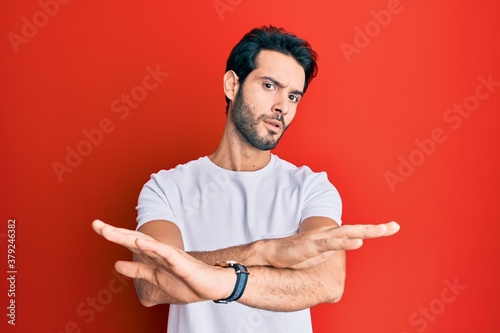 Young hispanic man wearing casual white tshirt rejection expression crossing arms and palms doing negative sign, angry face
