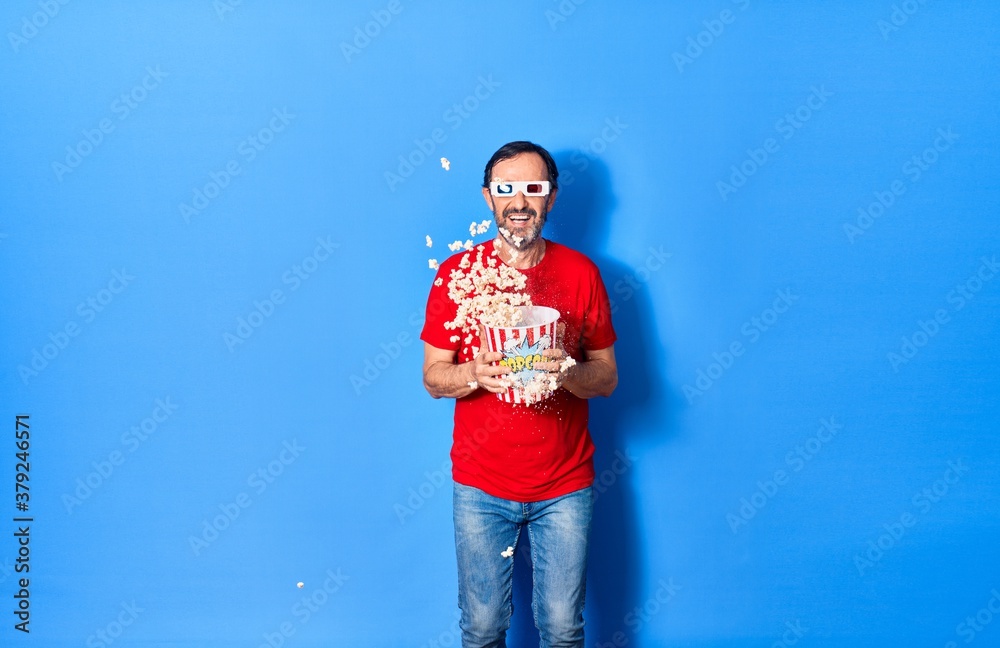 Middle age handsome hispanic man watching movie using 3d glasses similing happy. Holding bucket of popcorn jumping with smile on face over isolated blue background