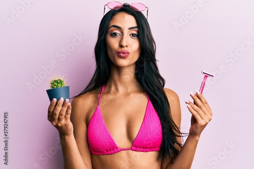 Φωτογραφία Beautiful hispanic woman wearing bikini holding cactus and razor looking at the camera blowing a kiss being lovely and sexy