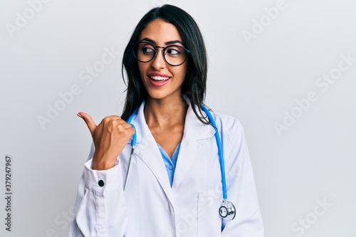 Beautiful hispanic woman wearing doctor uniform and stethoscope pointing thumb up to the side smiling happy with open mouth