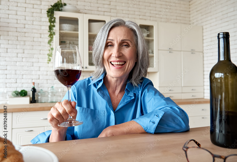 Foto Stock Happy 60s mature woman drinking wine video calling friend at  home. Old middle aged lady holding glass talking to web cam by online  social distance chat meeting sit at kitchen