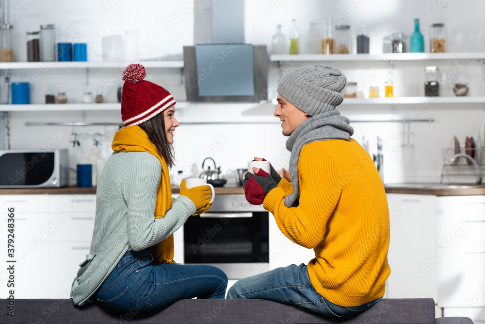 side view of young couple in warm hats, scarfs and gloves holding cups with tea in kitchen side view of young couple in warm hats, scarfs and gloves holding cups with tea in kitchen