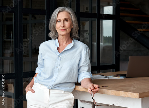 Confident stylish european mature middle aged woman standing at workplace. Stylish older senior businesswoman, 60s gray-haired lady executive leader manager looking at camera in office, portrait. photo
