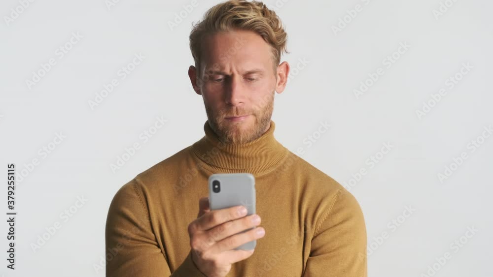 Attractive serious blond bearded man intently using smartphone making face grimace over white background Attractive serious blond bearded man intently using smartphone making face grimace over white background