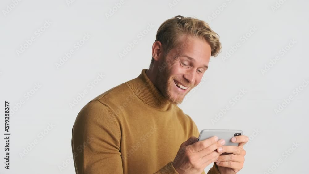 Attractive smiling blond bearded man happily watching video on smartphone over white background. Laughing expression Attractive smiling blond bearded man happily watching video on smartphone over white background. Laughing expression