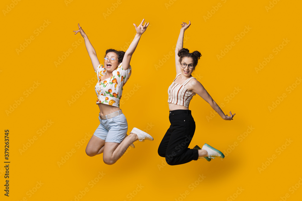 Caucasian twins jumping and gesturing happiness on a yellow studio wall while wearing summer clothes and glasses