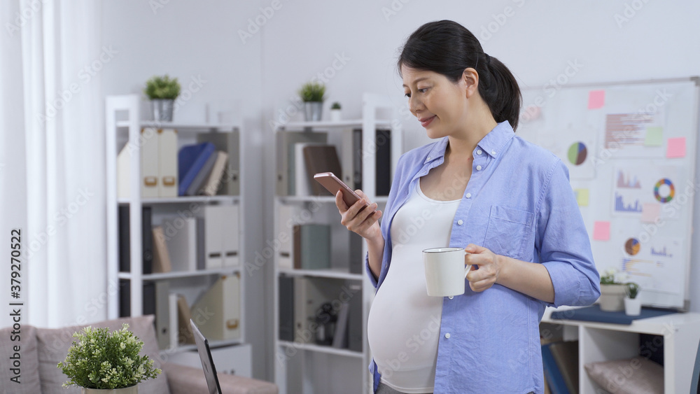 chinese pregnant female business owner in casual wear standing in the home studio with smartphone and cup of coffee.