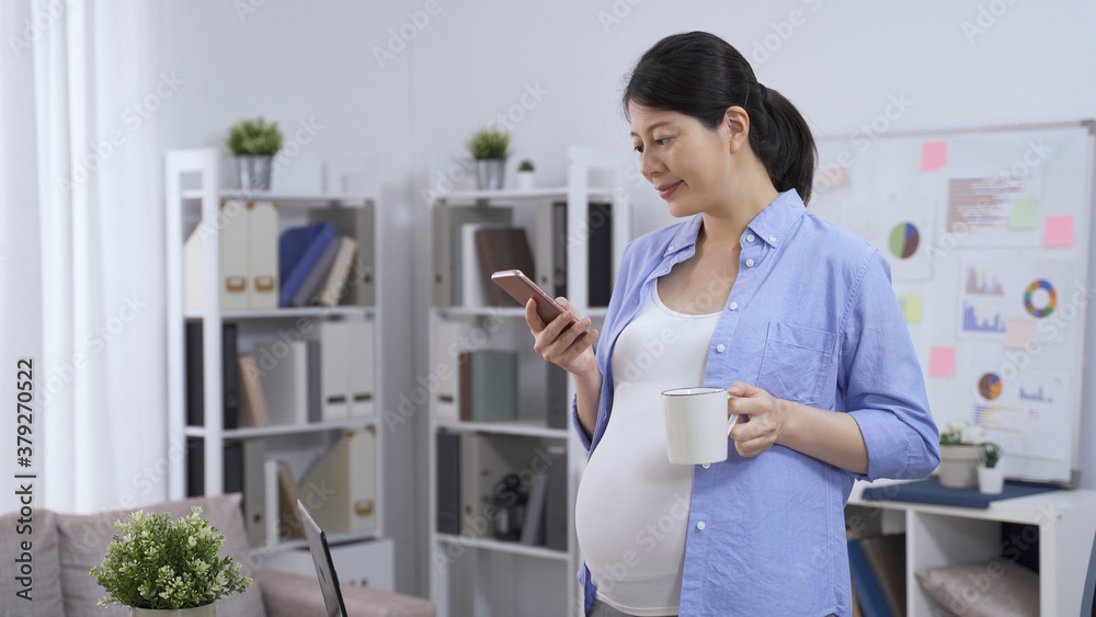 chinese pregnant female business owner in casual wear standing in the home studio with smartphone and cup of coffee. chinese pregnant female business owner in casual wear standing in the home studio with smartphone and cup of coffee.