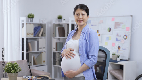 smiling asian korean female small business owner pregnant woman wearing light blue overshirt is gently touching and glancing at her swollen belly while looking at camera.