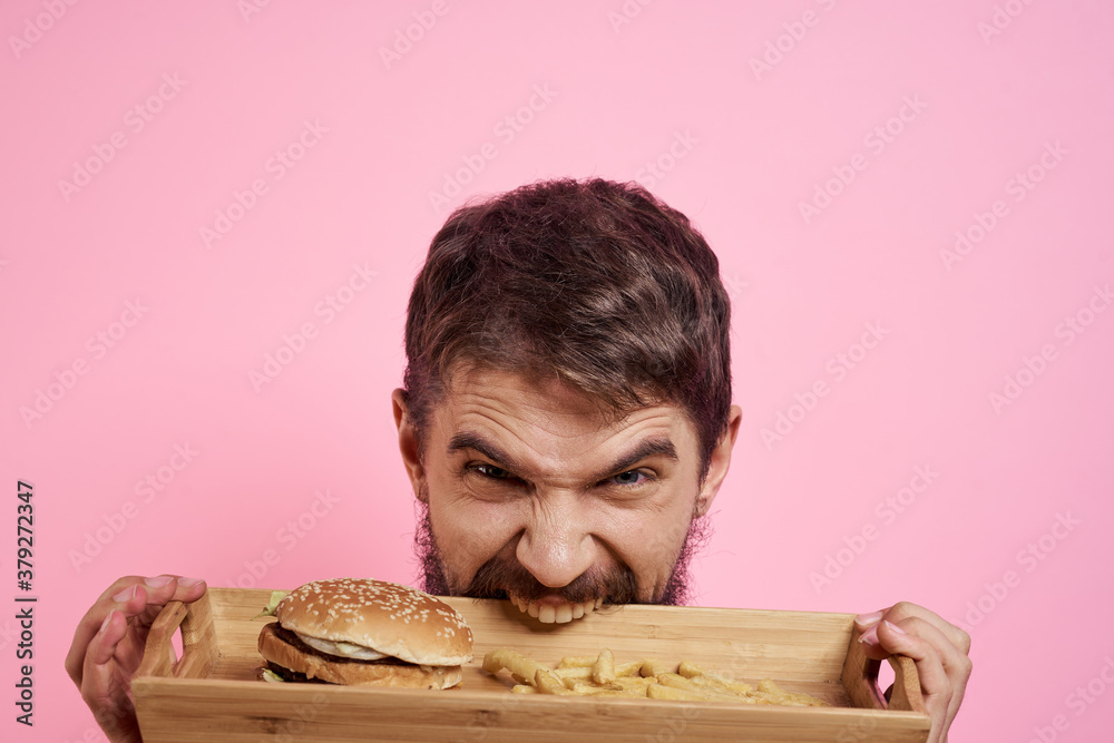 Aggressive guy with a tray in his hands hamburger fries pink background hungry look