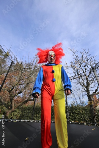 Halloween party outdoors.Scary clown with red hair jumping in a trampoline on autumn trees and blue sky background. Creepy clown costume. Festival and carnival. 