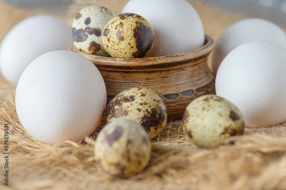 White raw chicken eggs and small quail eggs in a clay bowl on a burlap close-up. Chicken and quail eggs rustic background.