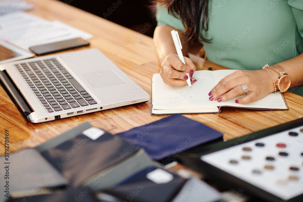 Fototapeta premium Close-up image of female atelier owner sitting at table with opened laptop and writing in planner