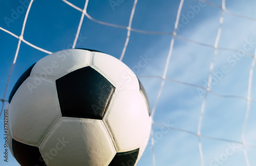 View of classic leather soccer ball in goal net on blue sky nature background. Traditional black and white football equipment to play competitive game sport tournament concept. 