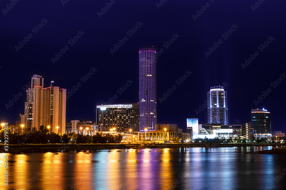 Obraz premium Cityscape with skyscrapers and other modern tower buildings with colorful illumination standing on the bank of river with reflections in it against dark blue sky at night. Horizontal orientation image