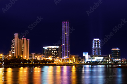 Cityscape with skyscrapers and other modern tower buildings with colorful illumination standing on the bank of river with reflections in it against dark blue sky at night. Horizontal orientation image