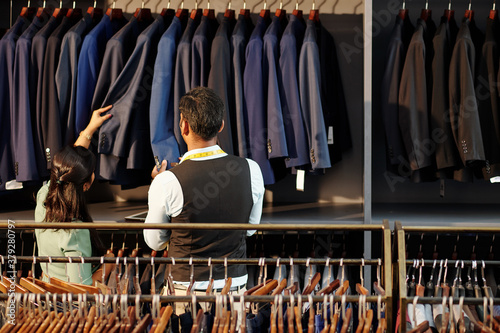 Head tailor and atelier manager checking quality of bespoke jackets hanging on rail, view from the back
