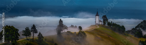 Jamnik, Slovenia - Foggy summer sunrise at Jamnik St.Primoz church. The fog gently goes by the small hilltop chapel on a wide panoramic photograph