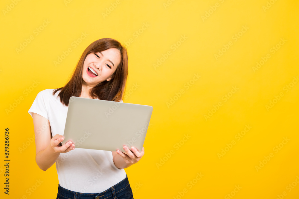 Asian happy portrait beautiful cute young woman teen smile standing wear t-shirt hold laptop computer and excited celebrating success looking to camera, studio shot yellow background with copy space Asian happy portrait beautiful cute young woman teen smile standing wear t-shirt hold laptop computer and excited celebrating success looking to camera, studio shot yellow background with copy space
