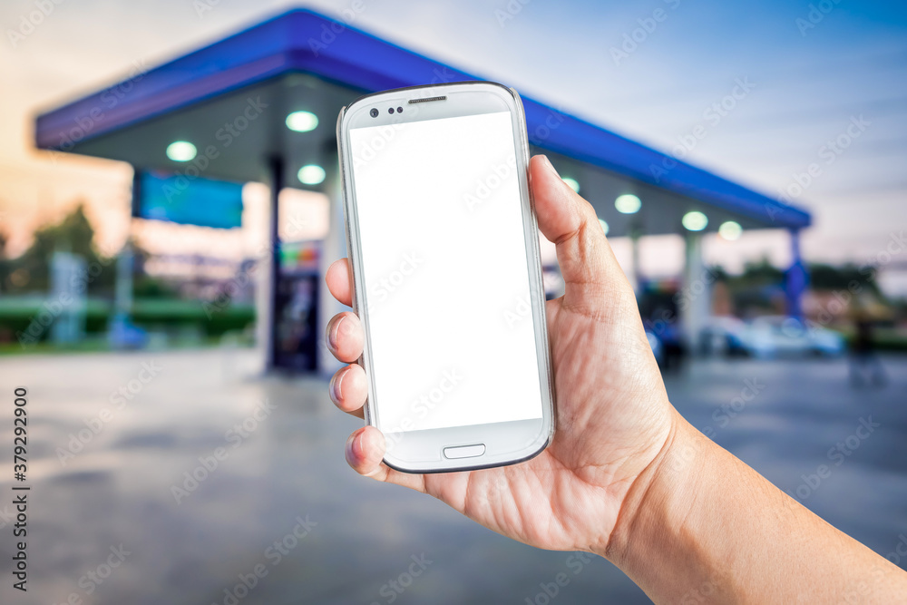 Hand holding a white smartphone with mockup blank screen blur background of twilight gas station at sunset. Touch screen mobile phone at the petrol station.