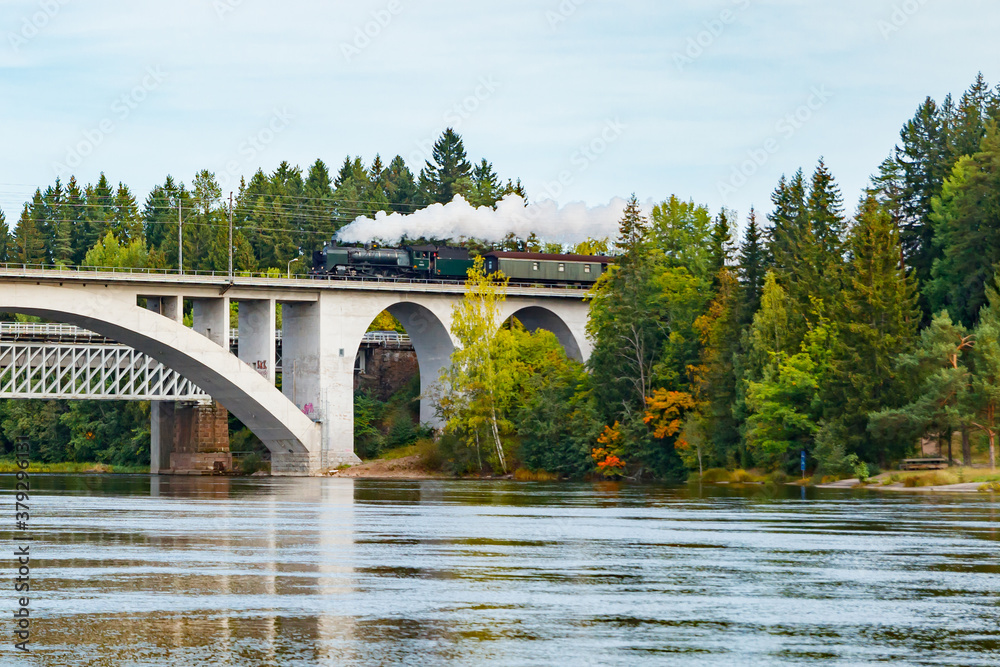 Fototapeta premium Kouvola, Finland - 18 September 2020: Autumn landscape of bridge with moving old steam passenger train Ukko-Pekka and Kymijoki river waters in Finland, Kouvola, Koria