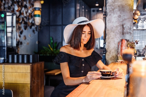 Beautiful, young woman of Caucasian nationality sitting at the bar and enjoying delicious morning coffee, cappuccino or americano. Dressed in a black dress and a white hat. Smiling cute
