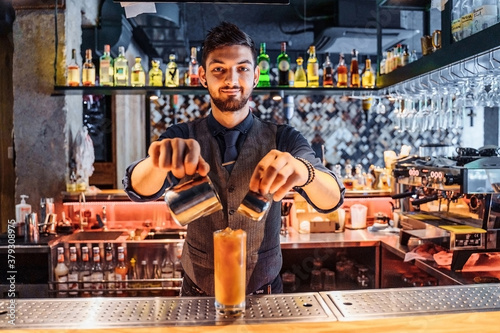 Handsome brunette barista of East Caucasian appearance preparing a drink at the bar in a restaurant. Mixing a coffee and orange juice by adding ice to the cocktail. Inserts a drinking straw