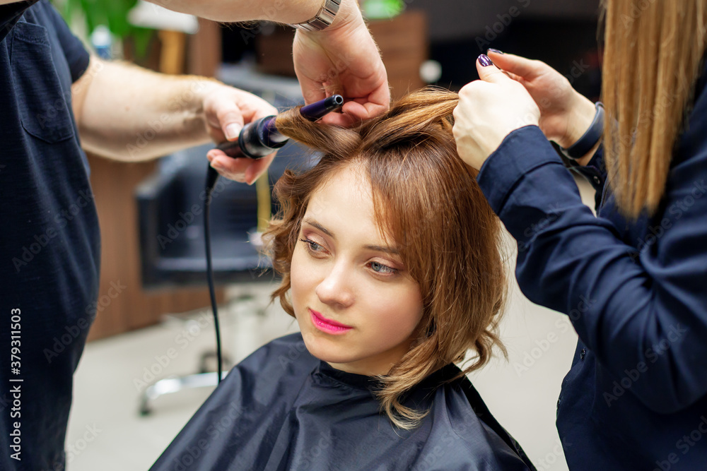 Professional hairdresser twists curls of long light brown hair of woman with curling iron in beauty salon. Hairdressing procedures Professional hairdresser twists curls of long light brown hair of woman with curling iron in beauty salon. Hairdressing procedures