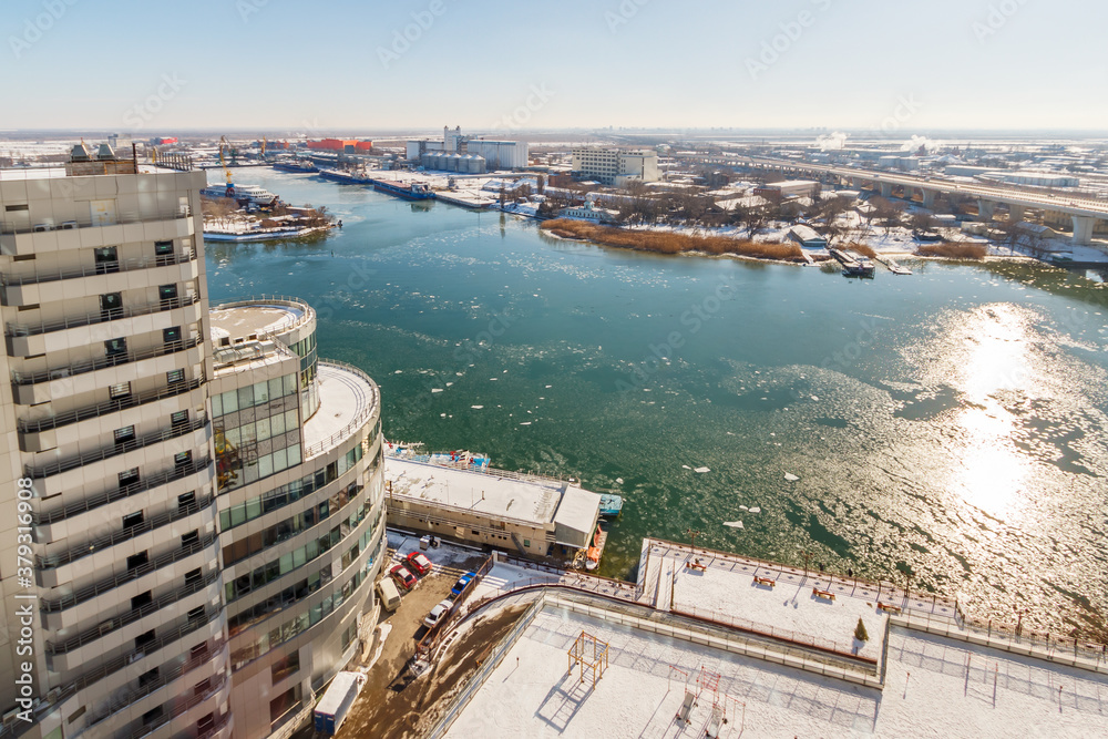 The sunny winter landscape with a river with floating ice and a high-rise residential building on the snowy riverbank