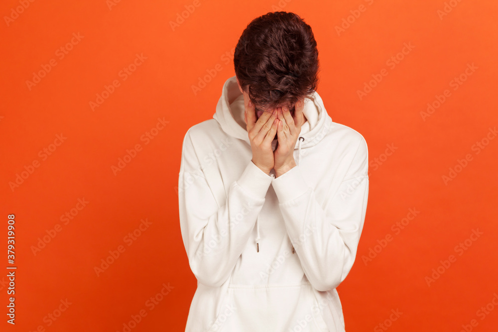 Upset lonely teenager in hoodie closing face with palms crying with despair, hiding his eyes in shame, depression. Indoor studio shot isolated on orange background Upset lonely teenager in hoodie closing face with palms crying with despair, hiding his eyes in shame, depression. Indoor studio shot isolated on orange background