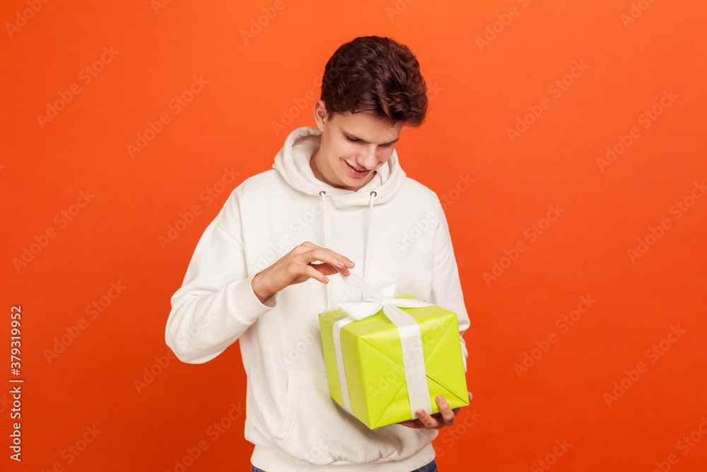 Handsome young man in casual white sweatshirt with hood adjusting bow on giftbox and smiling, teenager preparing for date, birthday gift. Indoor studio shot isolated on orange background Handsome young man in casual white sweatshirt with hood adjusting bow on giftbox and smiling, teenager preparing for date, birthday gift. Indoor studio shot isolated on orange background