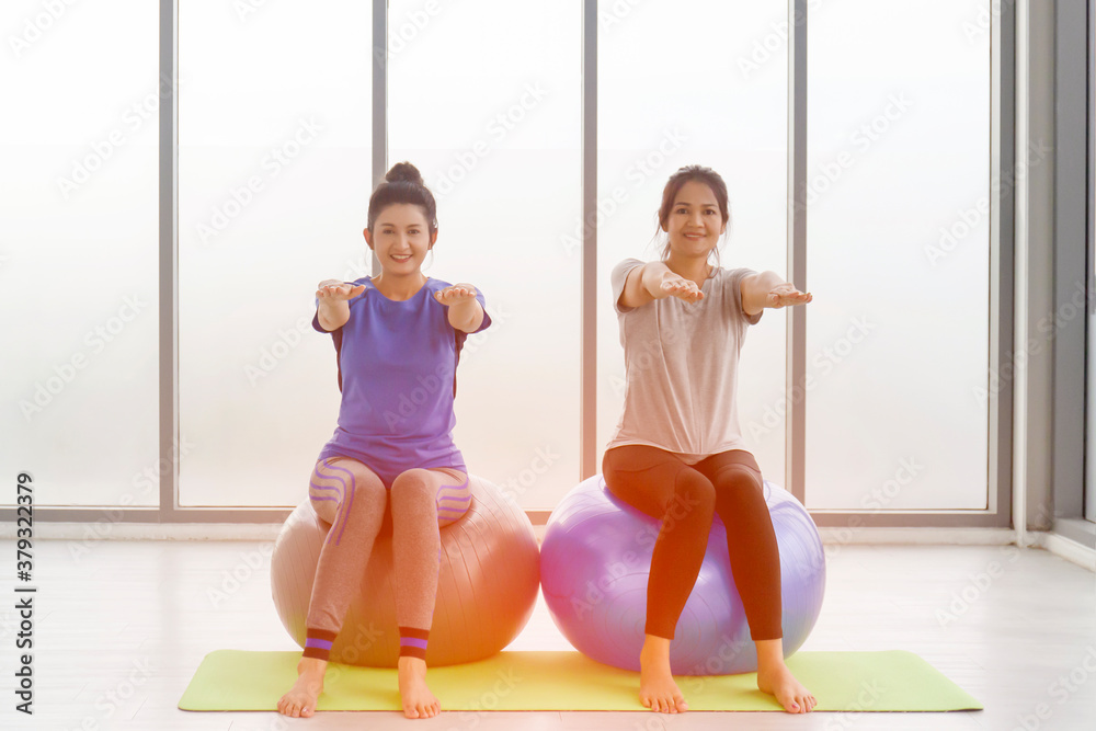 Naklejka premium Two middle-aged Asian women doing yoga sitting on a rubber ball in the gym.