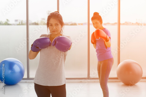 Two middle-aged Asian women are doing boxing exercises in the gym and have a light orange background.