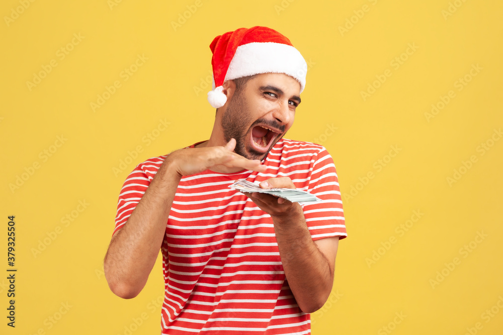 Extremely happy generous man in santa claus hat in red t-shirt making holidays gifts scattering dollars from hand, having fun. Indoor studio shot isolated on yellow background Extremely happy generous man in santa claus hat in red t-shirt making holidays gifts scattering dollars from hand, having fun. Indoor studio shot isolated on yellow background