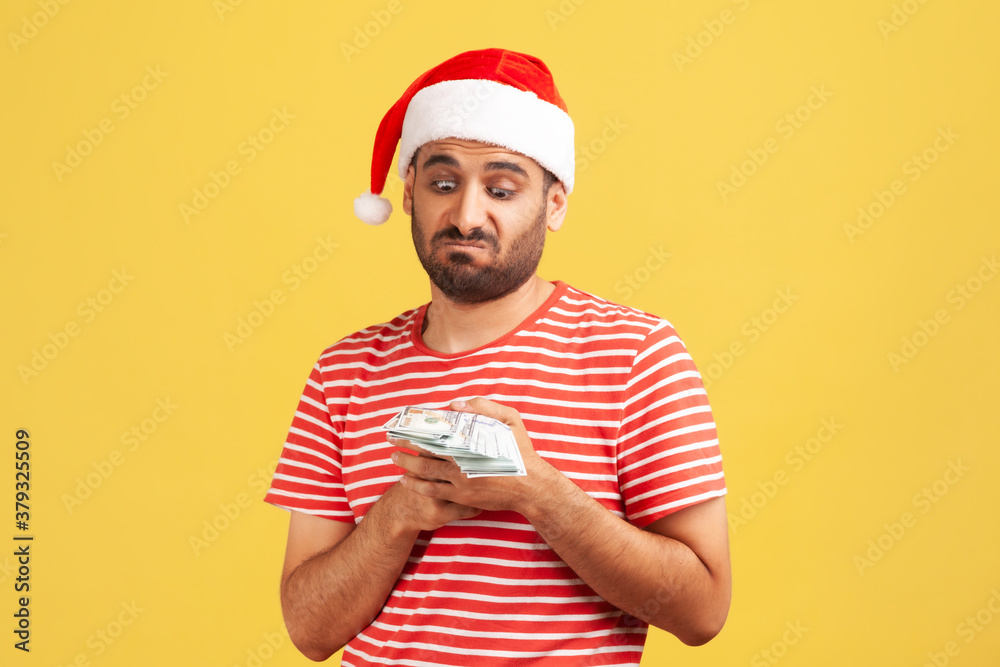 Unhappy confused bearded man in santa claus hat holding dollars in hands, greedy to buy holiday gifts. Indoor studio shot isolated on yellow background Unhappy confused bearded man in santa claus hat holding dollars in hands, greedy to buy holiday gifts. Indoor studio shot isolated on yellow background