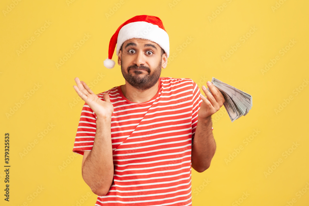 Puzzled confused man with beard in red santa claus hat holding cash in hands, dont know how to spend his allowance, holidays perks. Indoor studio shot isolated on yellow background Puzzled confused man with beard in red santa claus hat holding cash in hands, dont know how to spend his allowance, holidays perks. Indoor studio shot isolated on yellow background