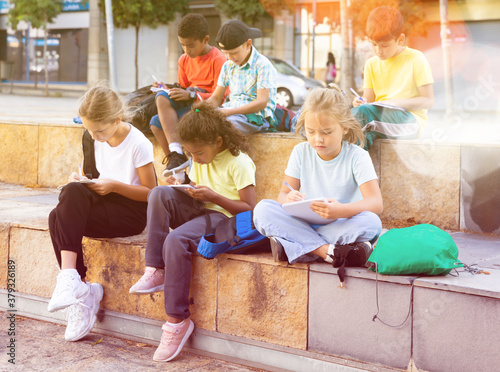 Multiethnic group of schoolchildren sitting with workbooks in schoolyard in warm autumn day.