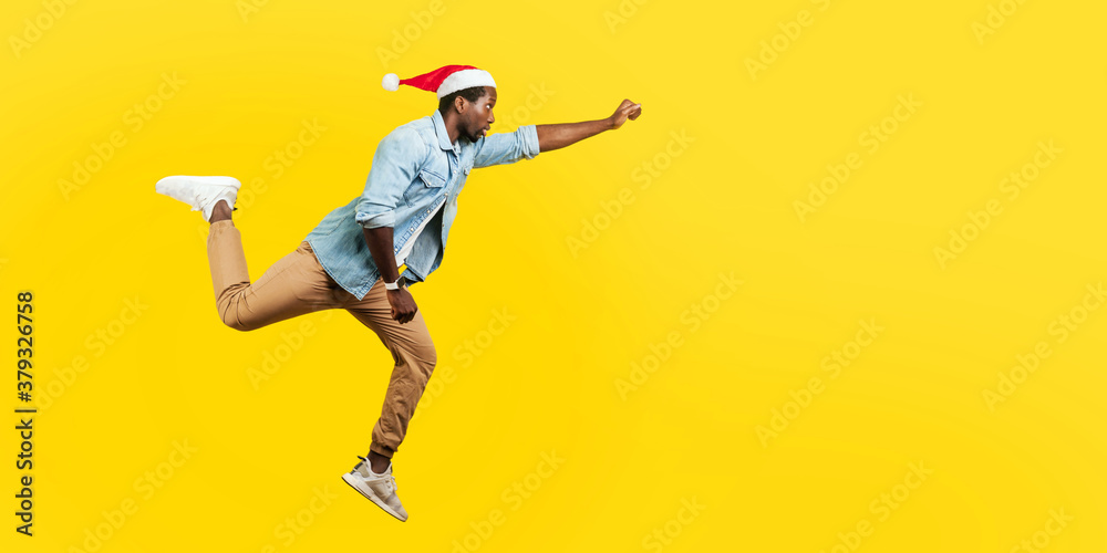 Full length portrait of handsome man in denim casual shirt and new year santa hat jumping in air or flying high with one stretched arm, feeling to be superman. indoor, isolated on yellow background Full length portrait of handsome man in denim casual shirt and new year santa hat jumping in air or flying high with one stretched arm, feeling to be superman. indoor, isolated on yellow background